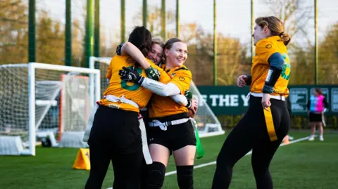 BAFA Four girls are hugging and celebrating while playing flag football. They are wearing black trousers and orange tops. They are on a pitch outside.