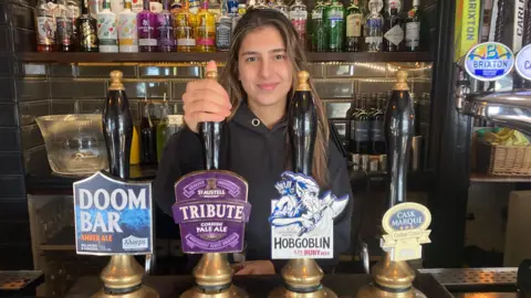 Pub owner Khara Schrijvers standing behind several pumps with the names of the different beers on display. 