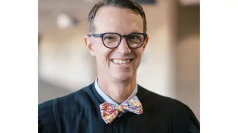 St Charles County Circuit Court A portrait of Judge Thornhill, showing him smiling. He has short black and grey hair, with black-framed round eyeglasses. He is wearing his judges robe and a colourful, patterned bowtie. 