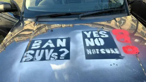 A close up of a range Rover car bonnet. There is stencilled grafitti on it with 'Ban SUVs?' and 'Yes No Not sure'. There are also red swirls daubed on the car too.