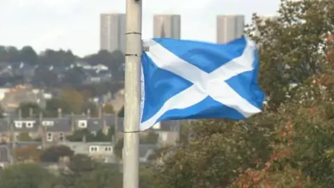 A blue and white saltire flag fluttering on a lamppost, with the city of Aberdeen in the background.