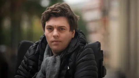 Emma Lynch/BBC Alex Taylor, a white man wearing a black jacket and grey scarf and in a wheelchair, on a London street