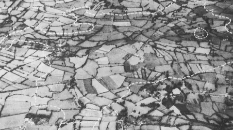 Bristol Water Old aerial black and white photograph. It has farmland, fields and trees. There is a white dotted line around where the Lake was proposed to go.