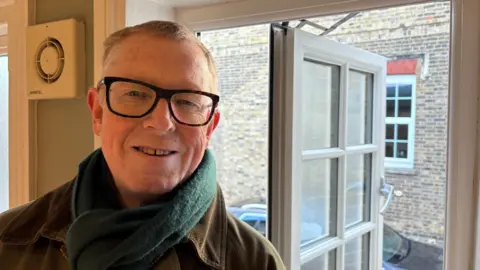 Martin Heath/BBC David Sanders with short brown hair, large black-framed glasses, wearing a green coat and green scarf. He is standing by an open window which looks out onto the brick wall of the neighbouring church.
