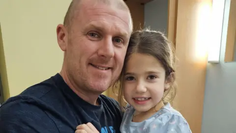Marie Wood A man wearing a blue t-shirt, holding his young daughter who is wearing a light blue pyjama top with a white star and moon print, both smiling at the camera.