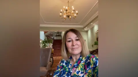 Ruth Richardson Ruth in a midshot in a large hotel-style foyer with a grand staircase behind her and a chandelier on the ceiling. She is wearing a bright multi-coloured shirt and is smiling to camera