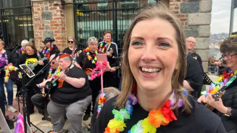 a woman smiles broadly. she is wearing a black t-shirt and a garland of mult coloured feathers and has light brown hair. behind her is a band of men and women dressed in a similar fashion, some stadning, some sitting. some are alos holding musical intruments