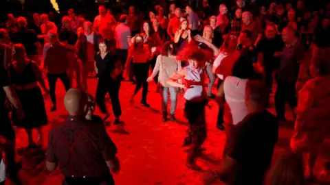 Around 50 people dancing in a room which is illuminated with red lights. 