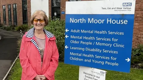 Tees, Esk and Wear Valleys NHS Trust Maureen Treharne is wearing a pink jacket with blue and white striped lining, and sunglasses. She is standing outside a building next to a huge blue sign for North Moor House, part of the TEWV NHS Foundation Trust