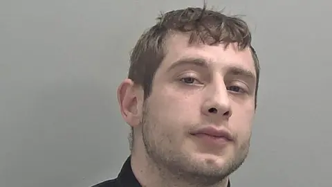 A mugshot of Oliver Beck. He has brown hair and slight facial hair and is wearing a black top.