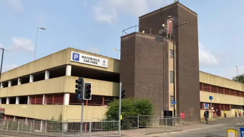 Alex Pope/BBC The outside of a large multi-storey car park, with a sign on it that reads "Mayorhold Car Park" and a person walking past. There are traffic lights in front of the building.