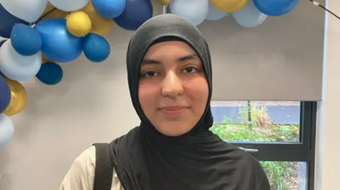 Charles Heslett/BBC A young woman in a hijab stands in front of a blue and yellow balloon arch.