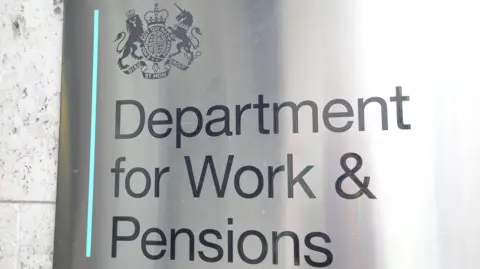 Close-up of a grey metal sign which says "Department for Work and Pensions". The government's logo containing a unicorn and a lion is above it in the top left corner. 