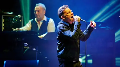 Getty Images James Prime, wearing a black waistcoat over a white shirt, is at a keyboard behind Ricky Ross who is singing into a microphone. He is wearing a dark jacket