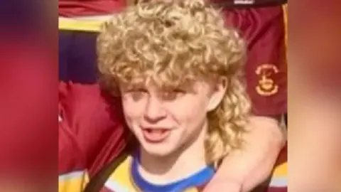 Malvern RFC Harry, a young boy with curly blonde hair, wearing a red and yellow top. Someone has their arm around him. It is a close-up from a group photo, and there are other people in the team kit around him.
