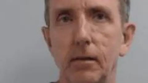 NCA A police mugshot of 57-year-old Charles Cornelius, who has grey hair swept to the side and is staring into the camera with a blank expression.