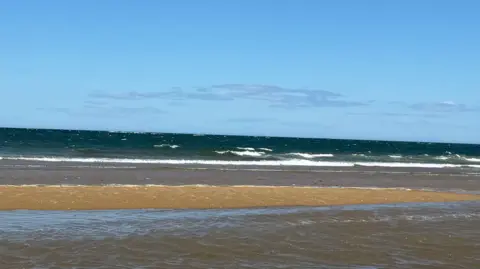 An empty stretch of the sandy beach at Redcar looking to the sea which is slighty frothed up by the waves. There is water pooled on the sand further inland.