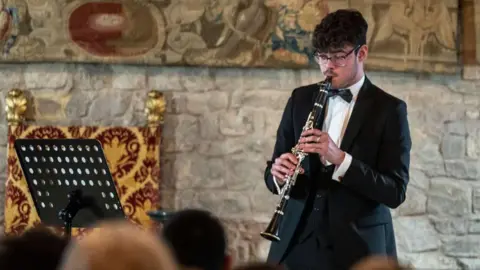 Malcolm Arnold Festival Christian Hoddinott, playing a clarinet, in a room, standing by a brick wall. A music stand is to the left. He is wearing a black dinner suit and has short dark hair, is wearing glasses and has a moustache. People are in front of him, in a blur.