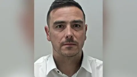 Custody image of Stephen Walkinshaw. He has parted dark brown hair, brown eyes and stubble. he is wearing an open-collared white shirt.