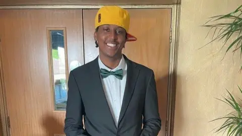 Eddie Kinuthia wearing a white collared shirt, green bow tie, black blazer and yellow cap styled sideways. He is standing in front of double wooden doors and smiling at the camera. 