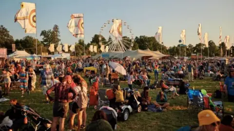 Mike Massaro A scene of people gathered at Wiltshire's Womad festival in 2024. Crowds are sitting and standing on the grassy site as colourful flags catch the wind, bathed in evening sunlight.