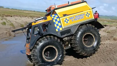 An amphibious large 4x4 vehicle that has huge wheels and looks like a small tank. It is black with blue and yellow emergency patterns on the side of the vehicle. It is travelling