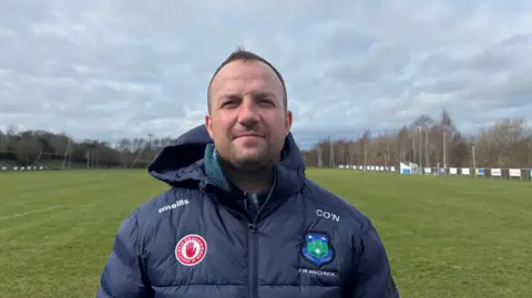 A man wearing a dark jacket with several sport badges on it. He has short brown hair and is looking at the camera. He is standing on a pitch. There are trees in the background. 