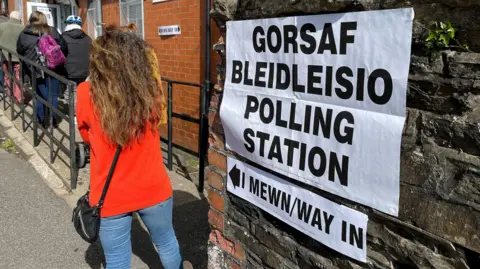 Getty Images The image shows voters waiting to enter a polling station. There is a bilingual notice on a stone wall in the foreground letting people know that they are at a polling station and how they should enter. It is a sunny day.