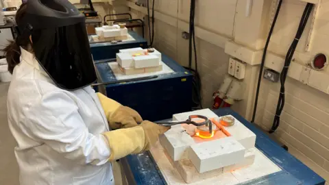 Kevin Church, BBC A scientist shows how nuclear waste can be 'baked' in solids like ceramic by making ceramic in a laboratory. The researcher wears a white coat and a protective helmet as it uses gloves and tongs to handle a shrine-thread of ceramic