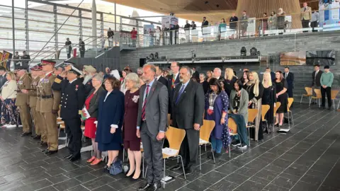 People standing in Senedd atrium wearing suits and formal clothing, with some in military uniform, looking solemn and some with heads bowed down