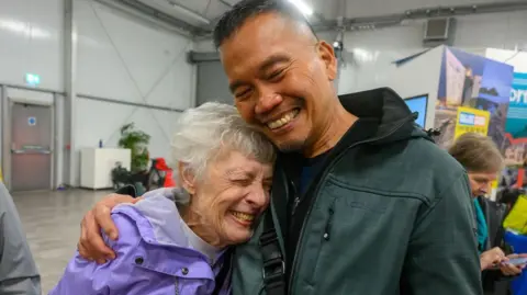 Pacemaker A woman with grey short hair in a purple raincoat is hugging a man with short brown hair in a green hoody. They are both smiling with their eyes closed. 