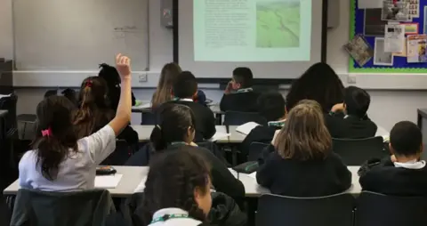 Stock image of the backs of children in a classroom. Rows of desks are in front of a projector screen, and among the children in uniform, one pupil raises their arm to answer a question
