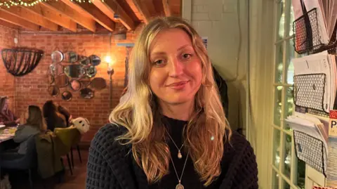 Holly Nichols/BBC A person, Georgia Chick, with long blonde hair sat indoors at a cafe with warm lighting. They are wearing a dark knitted sweater and a necklace with a round pendant. Behind them is a brick wall decorated with hanging mirrors and lights, and several people are seated at tables in the background. To the right, there are shelves holding books, papers, and baskets.