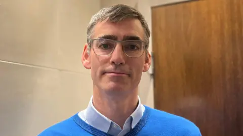 BBC Monty Tadier has short greying hair and is wearing glasses, and a blue jumper with a white shirt underneath.