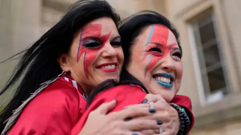 Two David Bowie fans, both with black hair and both with a red and blue lightning flash painted on their faces, hug outside the David Bowie World Fan Convention