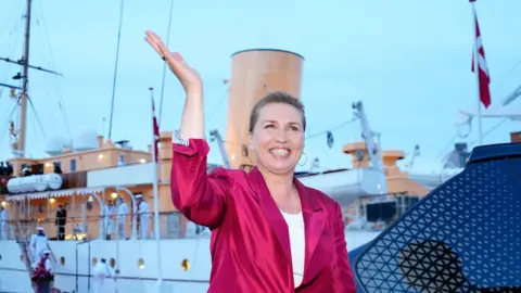 EPA Danish Prime Minister Mette Frederiksen waves, smiling, in a bright pink blazer. She stands in front of the royal yacht Dannebrog. 