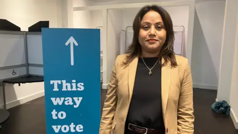 Amy Holmes/BBC A woman standing indoors beside a blue sign reading “This way to vote” with an upward arrow and “Milton Keynes City Council” at the bottom.