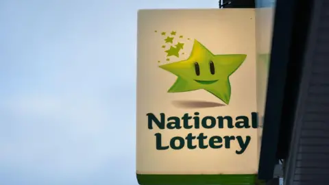 An Irish lottery sign on the side of a building. It has an image of an anthropomorphic green star with eyes and a smile, dancing along above the words "National Lottery" in a darker green. The whole sign has a white background and is illuminated by a light from within.
