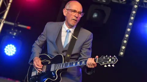 C Brandon/Redferns A man with short grey hair and brown glasses performing on stage. He is wearing a grey three-piece suit with a white shirt and light blue tie. He is holding and playing a black guitar