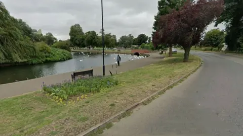 The River Nene as it curves round to the left, with a promenade pavement, strip of lawn, and road all following the same curve. There are swans on the river in the distance and you can see a small brick bridge that runs alongside the river. There are large trees either side of the river and a playground in the distance.