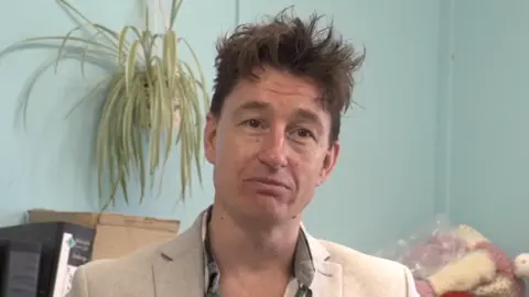 A man in a beige suit sitting in front of a light blue wall. He has brown hair.