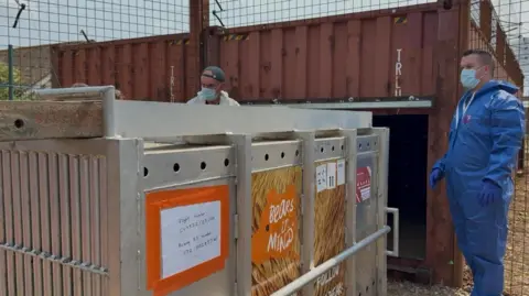 A large silver crate with a flight number printed on the side, with two mean wearing overalls and face masks standing next to it.