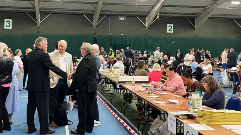 BBC Inside a hall during the election count. People are sitting at tables for the count.