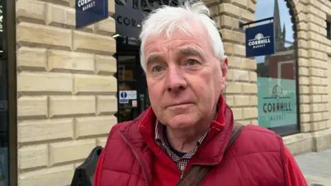 A grey haired man wearing a red gilet over a red fleece looks at the camera. He is standing in front of a large, lightly-coloured stone building which has large windows. There is a sign which reads Cornhill Market.