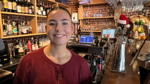 Interior of a bar with shelves stocked with bottles of wine and spirits against a brick wall. A wooden counter in the foreground holds cocktail-making equipment, a metal shaker, and fruit. Hanging glassware is visible above the bar, and a computer screen is on the back counter. A Santa hat is placed on a beer tap, adding a festive touch.