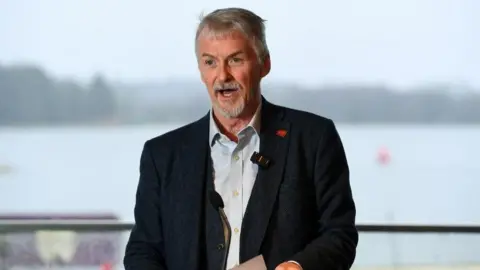 Welsh government Huw stands on a podium with a body of water behind him. His mouth is open with a word and he is holding a paper in both hands He has a dark suit jacket and light blue shirt. He as short grey hair 