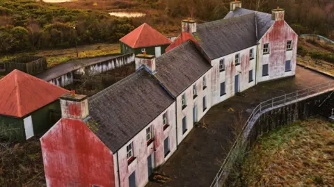 Jay Curtis Close up drone shot of a street in the village. The houses are white with black bricked roofs. They are derelict with red marks on their sides.
