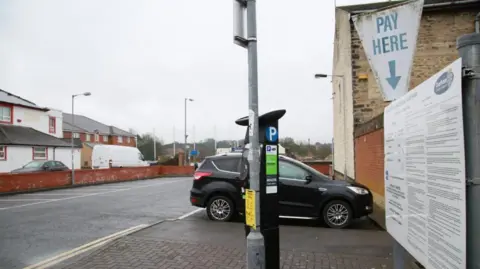 A black car is parked outside on a paved park off-road on a residential street. There is a black parking meter behind a lamppost. In front of a wall there is a sign listed by Durham County Council with a list of information. 