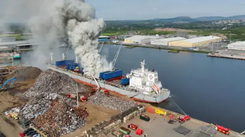 A drone shot of the Nord Houstong in Newport Docks. White smoke is billowing from the centre of the ship, which is docked next to huge piles of scrap metal. Large docks buildings can be seen across the water in the background and fire engines are stationed close to the ship