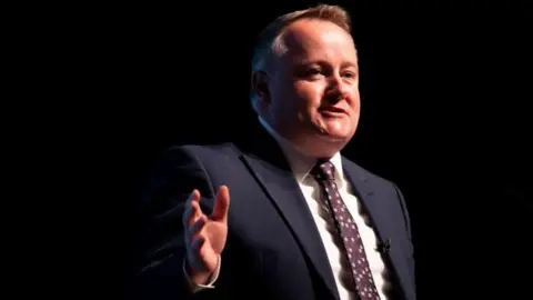 Darren Millar gesturing away from himself, stood on a stage. Only his upper body is visible and he is wearing a dark suit, tie and white shirt.